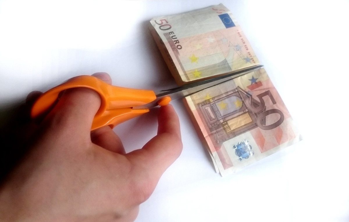 A hand cutting a fifty-euro note in half with orange scissors.